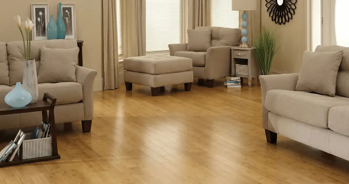 A living room with professionally installed bamboo flooring, beige sofas, a matching ottoman, a side table with a blue lamp, vases, framed pictures, and large windows with blinds and curtains in Jacksonville, FL